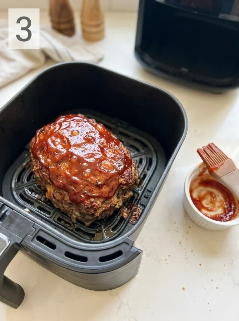 Browned air fryer meatloaf topped with ketchup glaze, beside a sauce bowl and pastry brush.
