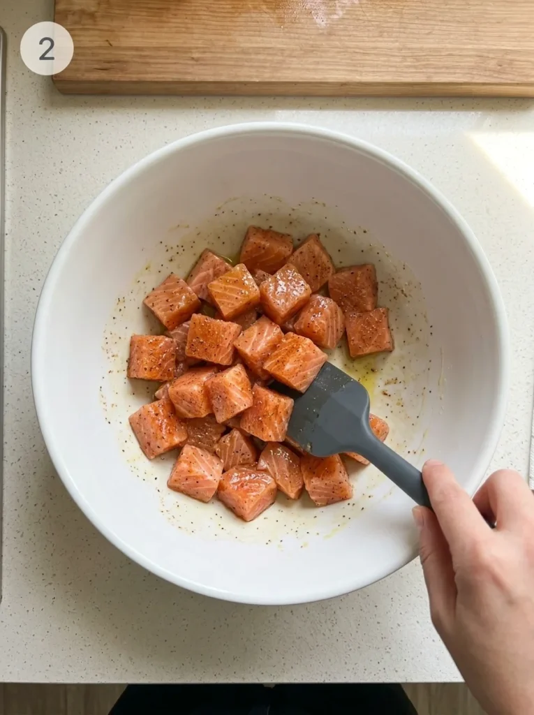 Raw salmon cubes being mixed with olive oil, garlic powder, and smoked paprika in a white bowl